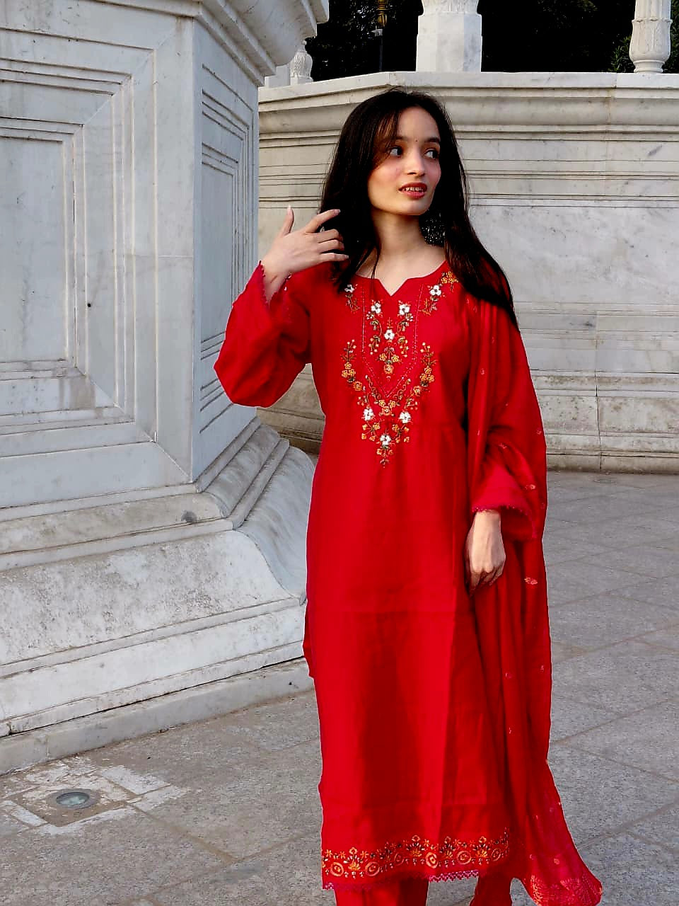 A vibrant red ethnic suit named Noor, showcasing elegant hand-embroidery and a scalloped border, styled for a festive look in a natural outdoor setting.
