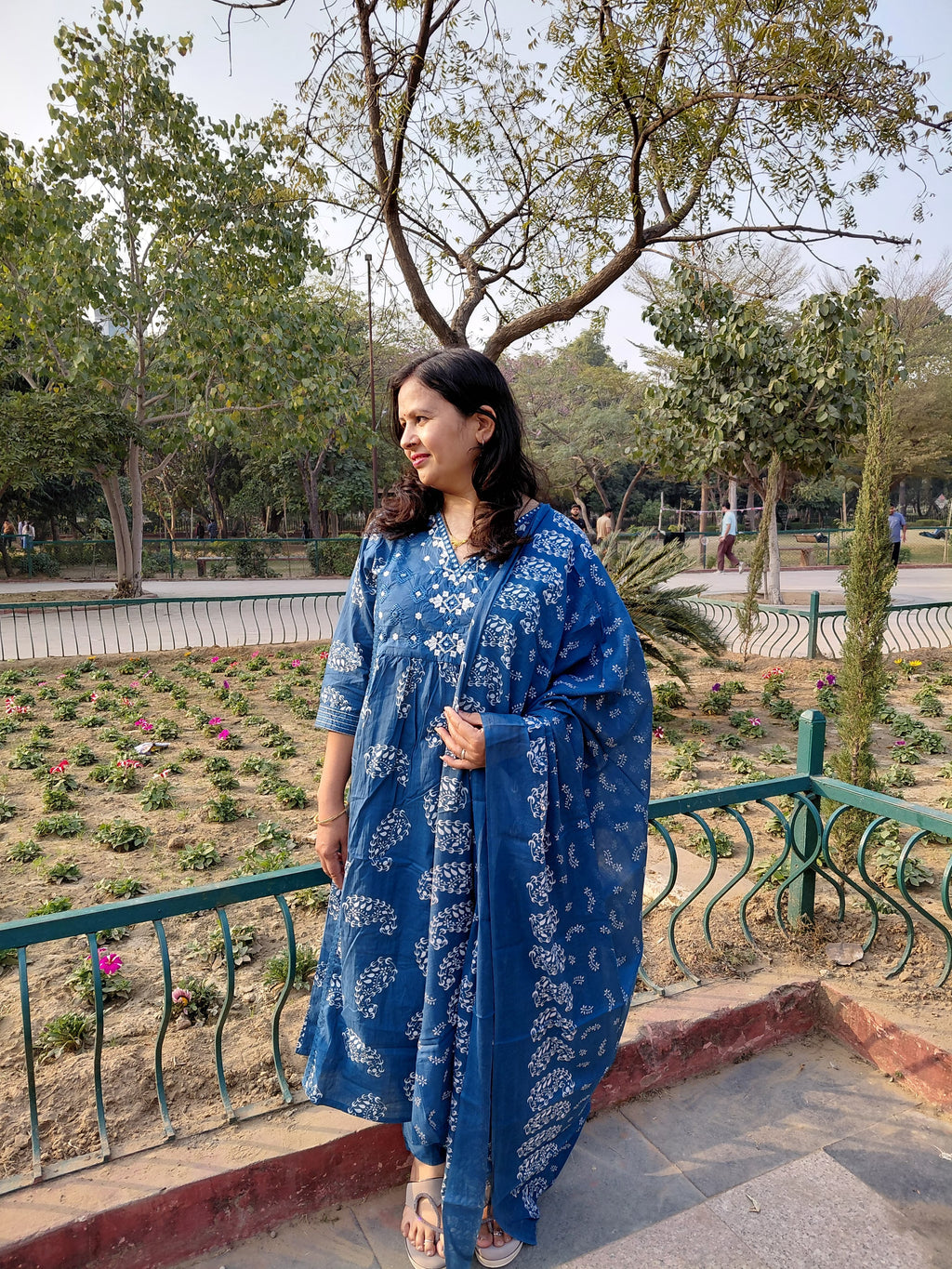 A woman in The Reet Set | Indigo Block Print stands near a flower bed in a park, looking to her right with trees and greenery in the background.