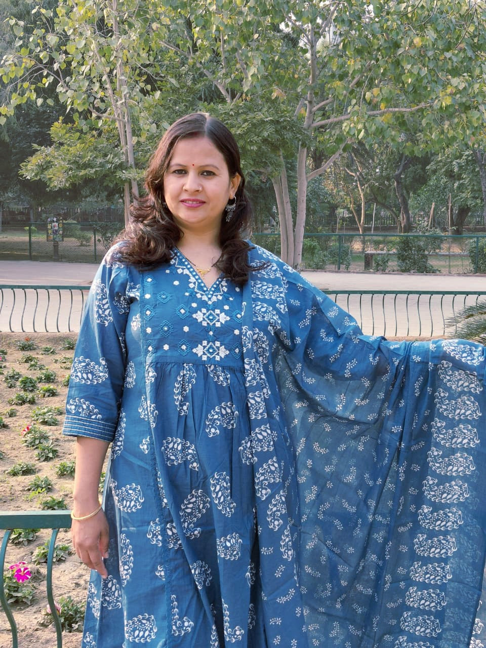 A woman in The Reet Set | Indigo Block print Anarkali poses gracefully outdoors, holding her dupatta amid a serene garden with lush greenery and a metal fence.