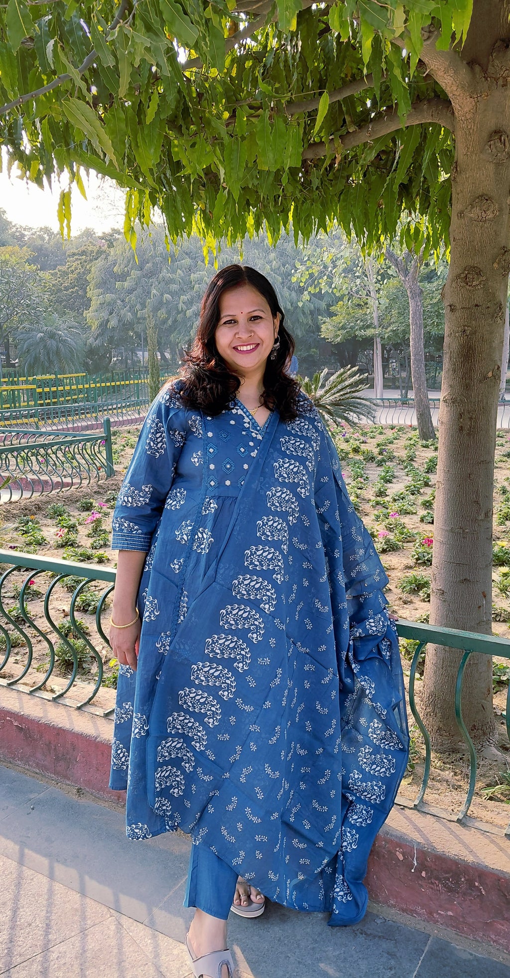 A woman in The Reet Set | Indigo Block print smiles by a green railing in a sunlit garden, surrounded by trees and flowerbeds.