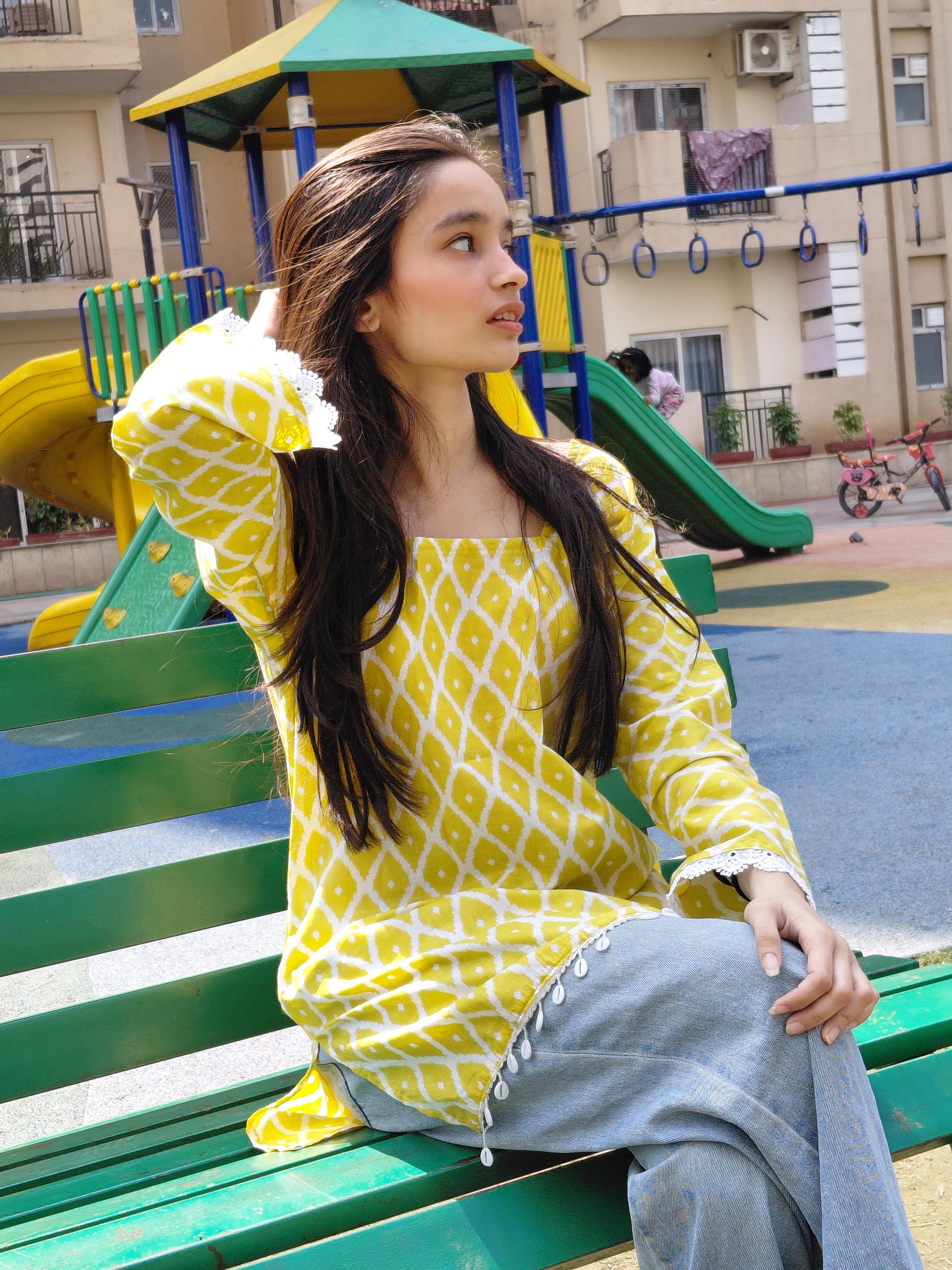 Woman in yellow patterned dress sitting on a green bench with a colorful playground in the background