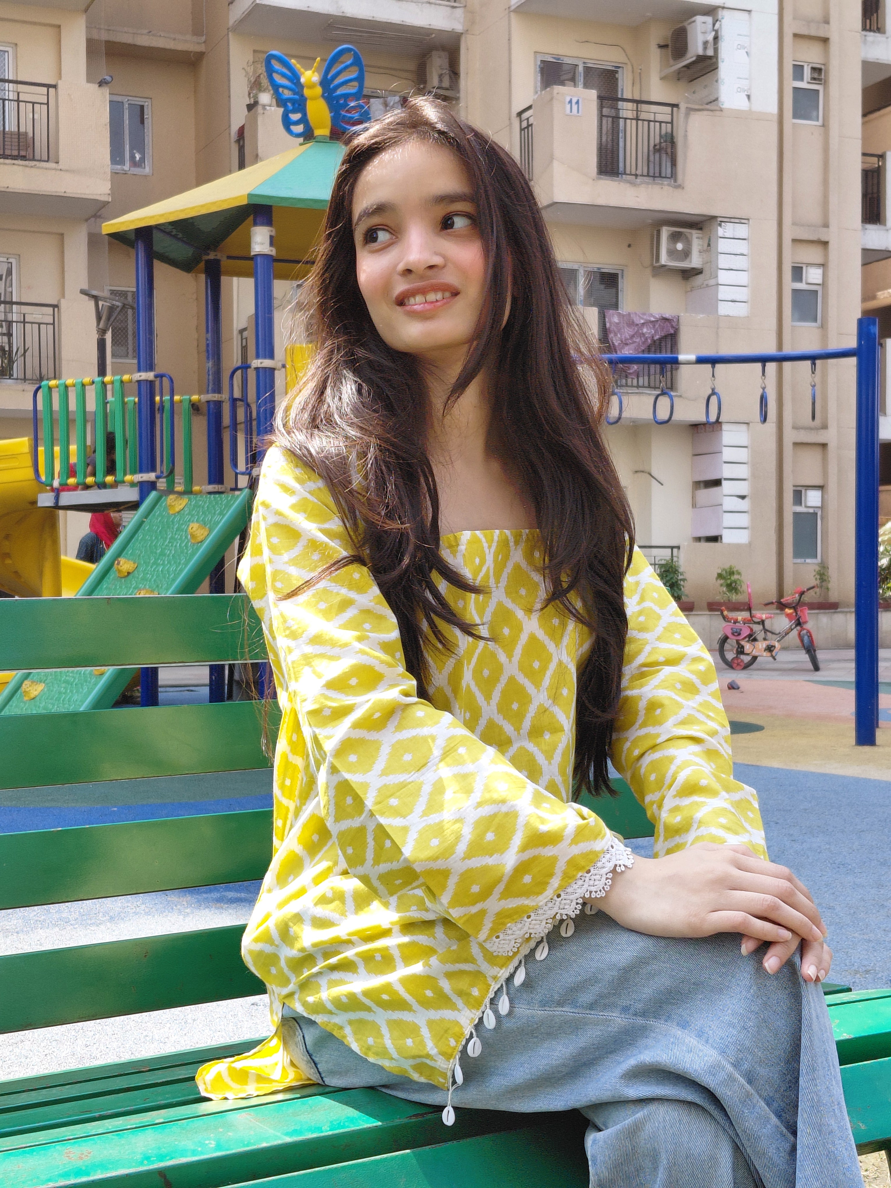 Person wearing a yellow patterned dress standing in front of a building with colorful playground equipment.